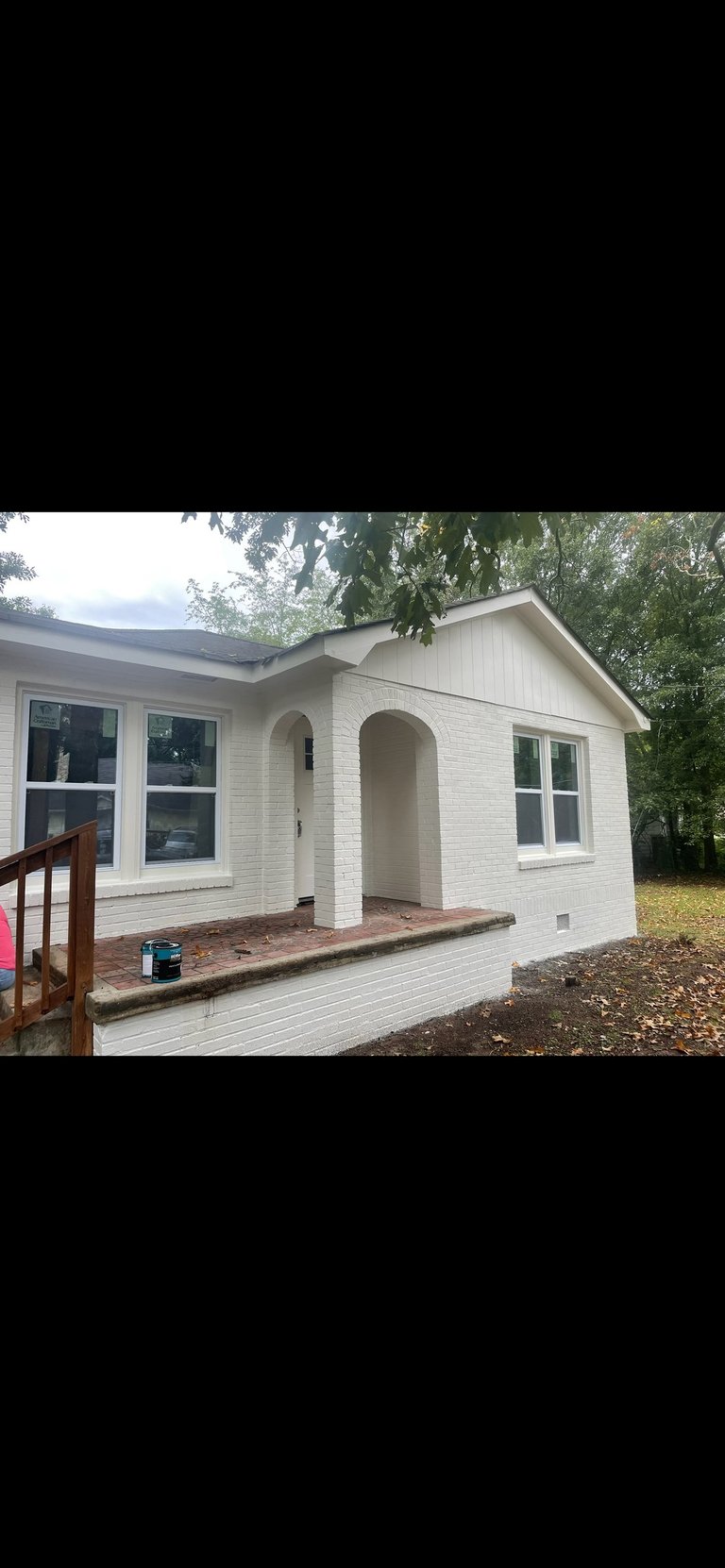 White single-story house with brick foundation, arched entryway, large windows, and front porch surrounded by trees