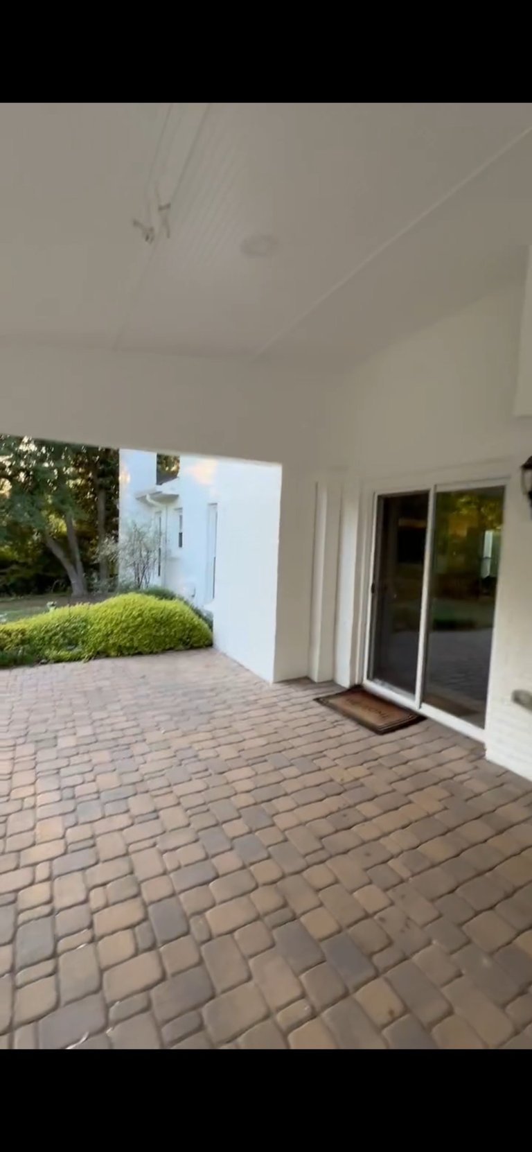 Covered patio with brick pavement leading to glass sliding doors of a modern white house, with manicured green hedges and white exterior wall visible