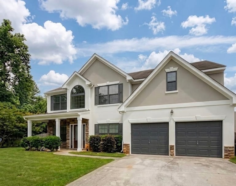 Modern two-story white house with two dark gray garage doors, manicured lawn, and blue sky with white clouds
