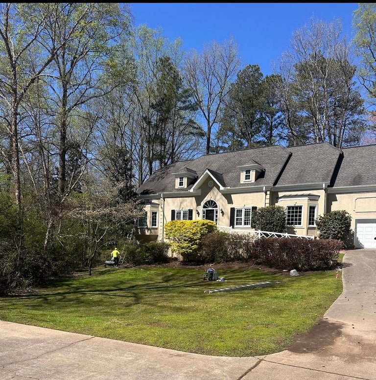Cream-colored two-story house with black roof and white shutters, surrounded by landscaped lawn and trees on a sunny day