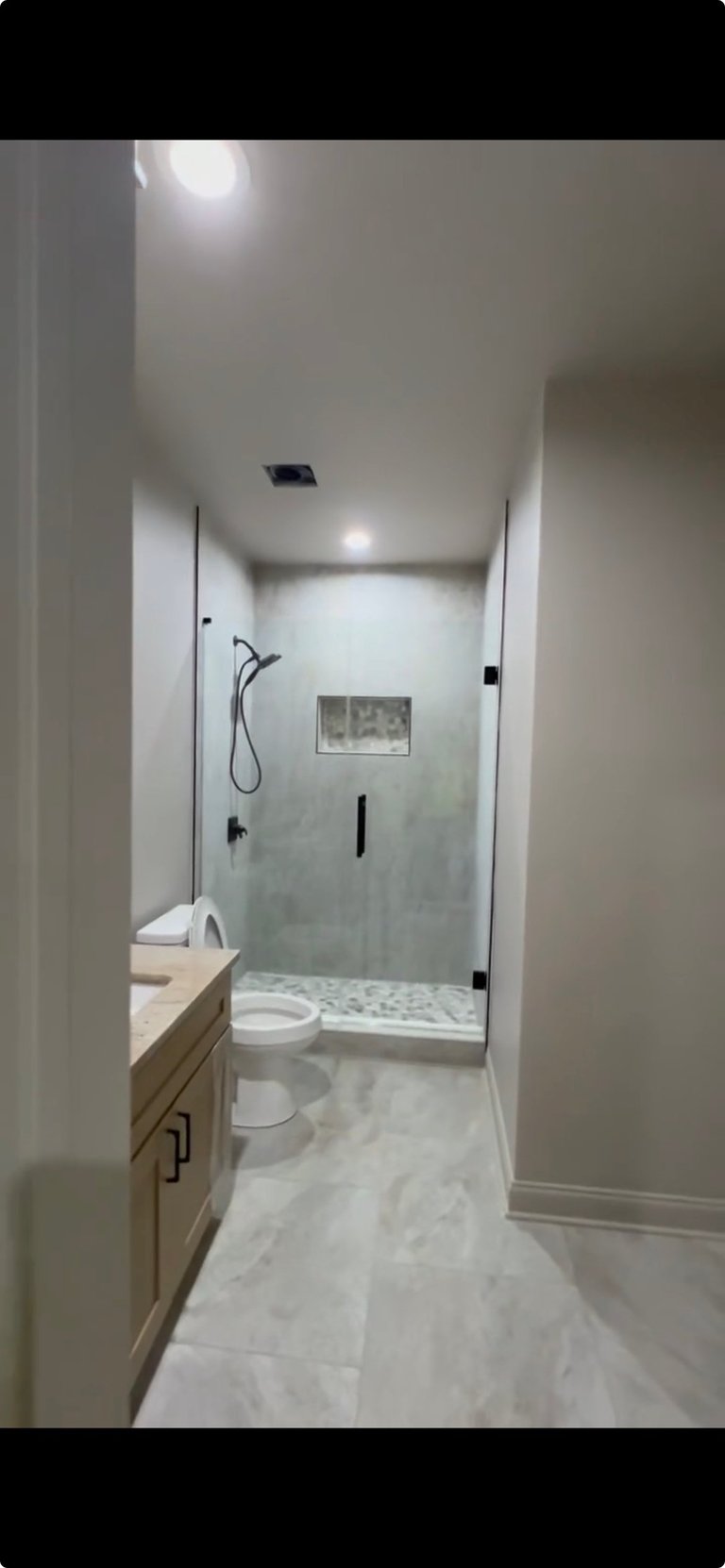 Modern bathroom with white vanity on left, glass-enclosed walk-in shower with gray tile on right, marble flooring, and recessed lighting