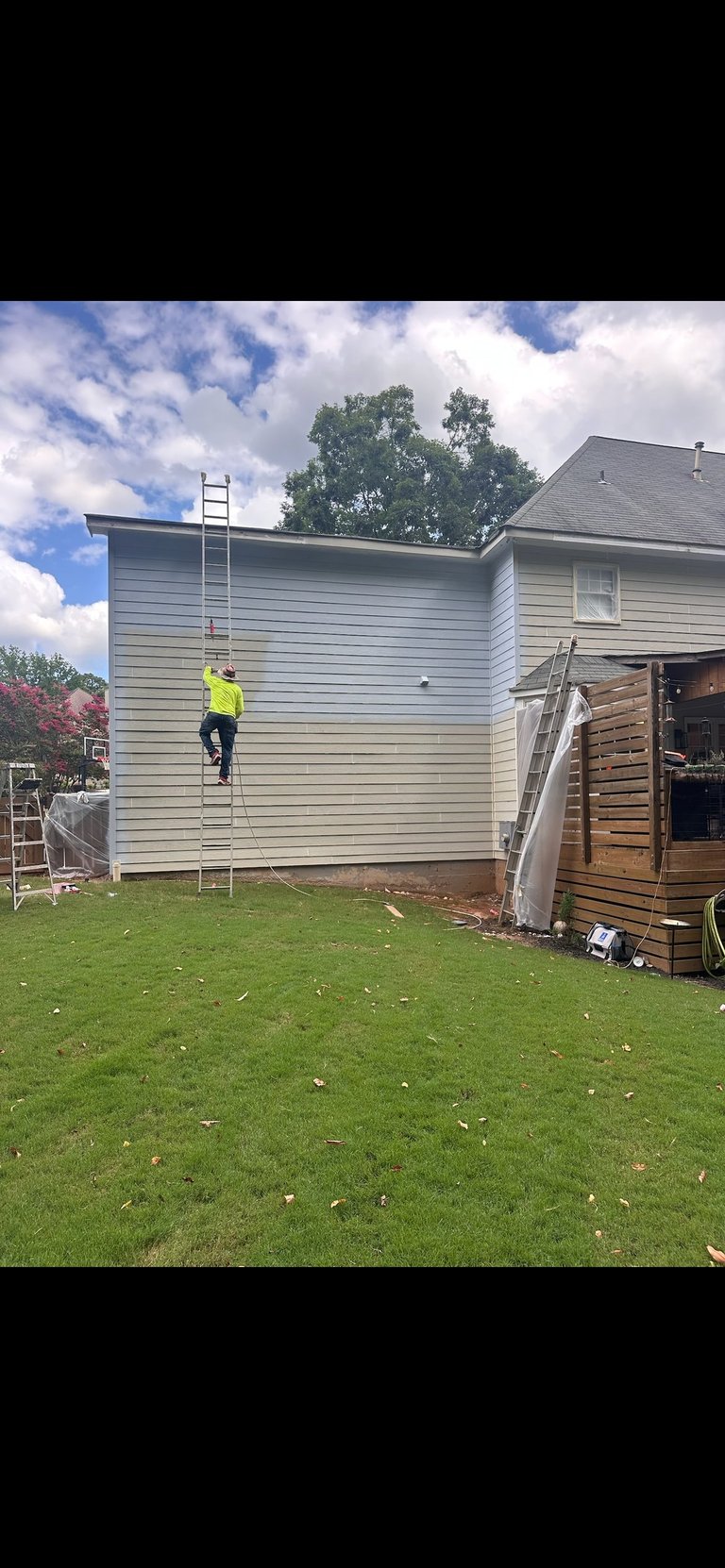 Worker in yellow safety vest climbing metal ladder on gray sided house addition during construction on green lawn