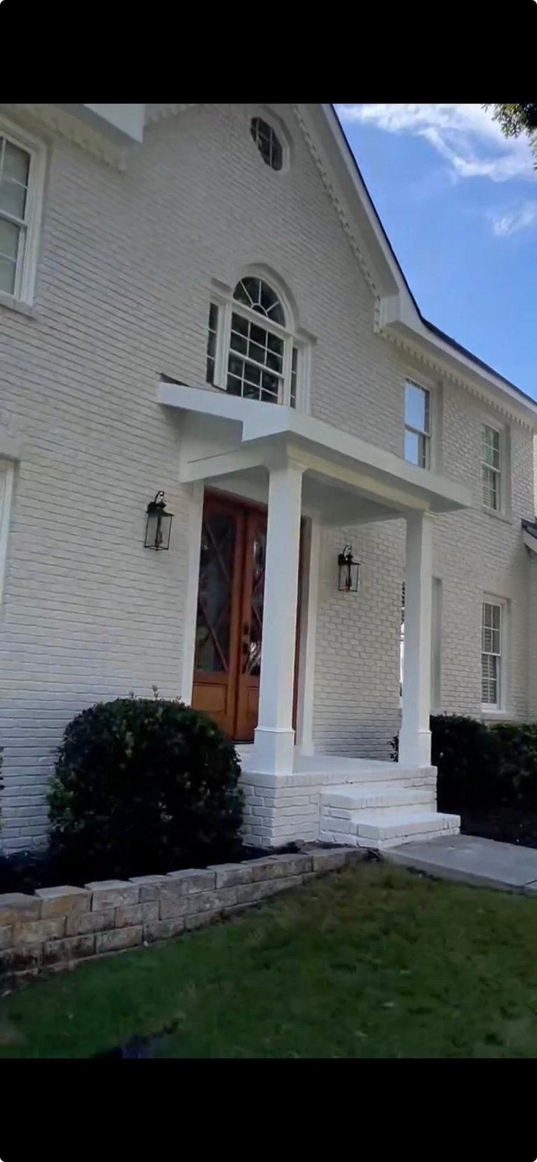 Front exterior of a white brick farmhouse with a covered porch entry, wooden red door, and manicured landscaping on a sunny day