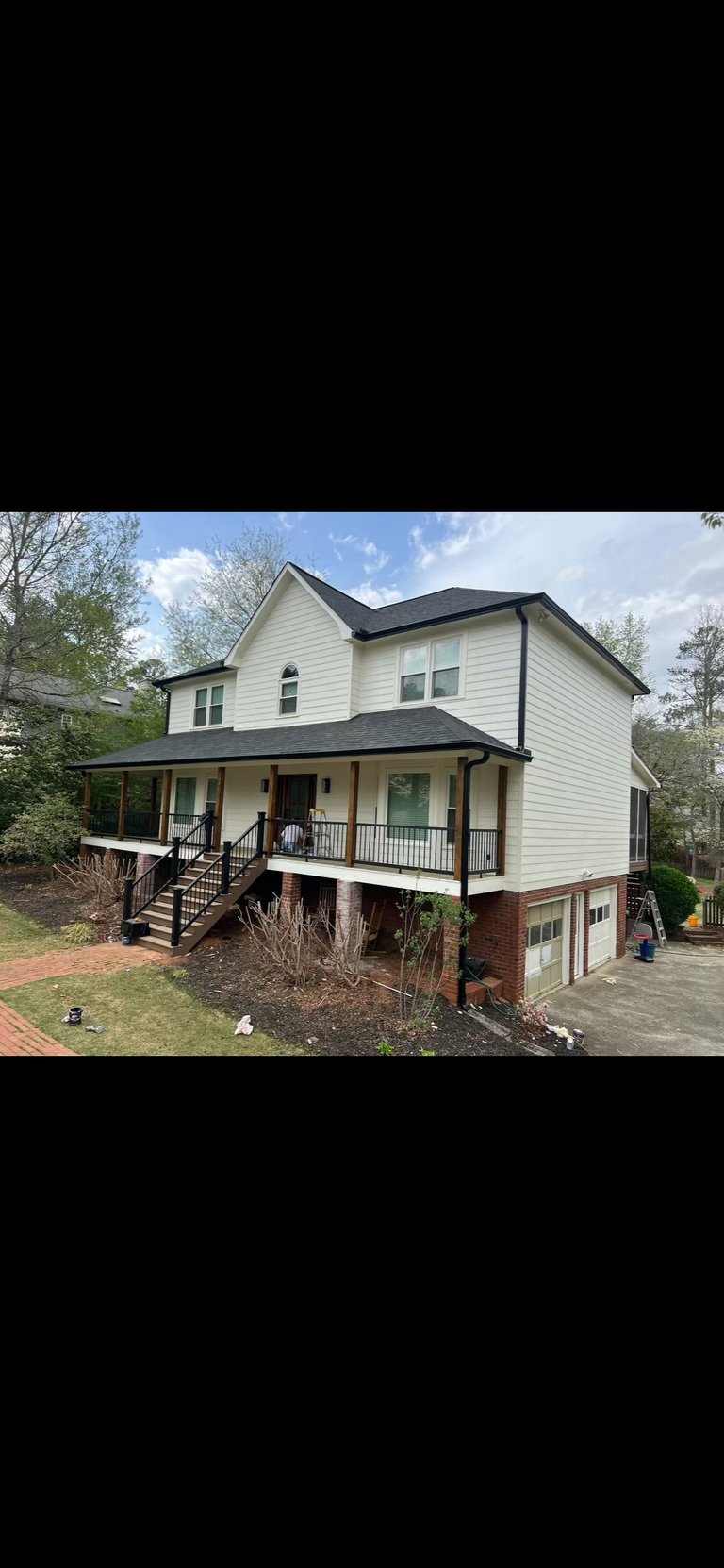 Two-story beige house with black roof, front porch, and attached garage surrounded by trees and landscaping