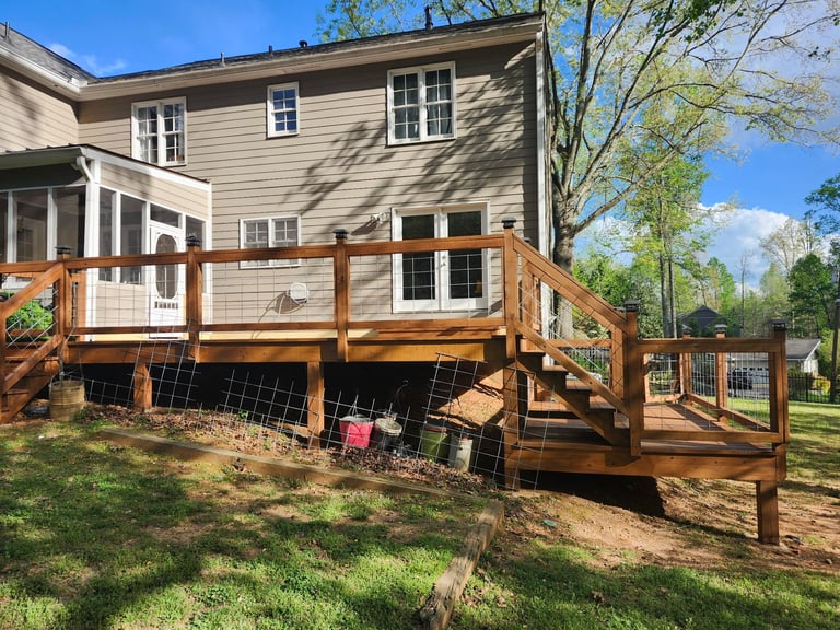 Beige two-story house with wooden deck and railing on a sunny day with green lawn and trees